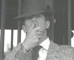 Black-and-white image of a man seen from mid-chest up, wearing a fedora and a jacket with a houndstooth-like pattern. He holds a cigarette between the middle and index fingers of his left hand and strokes his upper lip with his thumb. He stands in front of what appears to be a mirrored doorway.