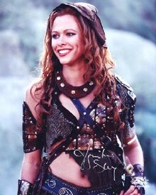 Upper body shot of a young woman with long curly brown hair, she is wearing leather bra, skirt and hat.