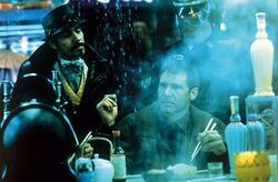 A man with close-cropped hair wearing a brown jacket sits at a counter, holding a pair of chopsticks poised over a rice bowl. Rain cascades down beside him as if from the edge of an awning. In the foreground is a teapot, several bottles, and other dining accessories. Steam or smoke rises from an unseen source. In the background, two standing men look down at the central figure. The goateed man on the left wears a dark snap-brim hat, a black coat with upturned collar, and a gold-trimmed vest. The man on the right, partly obscured by the steam, is wearing a constabulary-style uniform, featuring large wrap-around shades and a hat or helmet with a glossy, stiff brim. There is a bluish cast to the entire image.