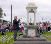 Photo of an ANZAC memorial with an elderly man playing a bugle. Rows of people are seated behind the memorial. Many small white crosses with red poppies have been stuck into the lawn in rows on either side of the memorial.