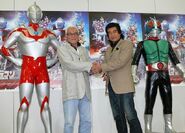 Susumu Kurobe holding hands with Hiroshi Fujioka next to the statues of their respective henshin forms.