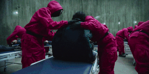 Workers change clothes of the unconscious players while Kang Sae-byeok conceals her switchblade.