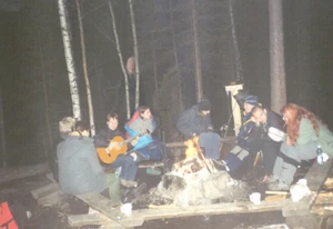 Slender Man standing in the trees of a forest at a campsite.