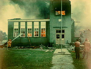 Slender Man standing on top of a building while it is on fire, in the smoke..