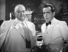 Black-and-white film screenshot of two men, both wearing suits. The man on the left is older and is nearly bald; the man on the right has black hair. In the background several bottles of alcohol can be seen.