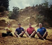 The Wiggles stiting in a sandy area