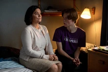 Queen Kristina sits upright in a beige skirt, sweater, and blouse. Next to Wilhelm, who is in a purple New York City Opera t-shirt. She is not looking at him, but he is mid exclamation towards her.